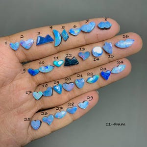 Puede incluir: Una colección de ópalos en diversas formas, incluyendo corazones y óvalos, mostrando una gama de colores, principalmente azules y verdes. Algunos ópalos exhiben cualidades iridiscentes. Tamaños de 4mm a 11mm.
