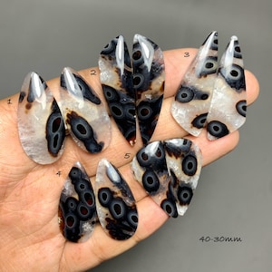 May include: A collection of ten polished, teardrop-shaped cabochons featuring a translucent white base with distinct black and brown circular eye-like patterns. The stones are numbered one through five, with a size range of 30-40mm indicated.