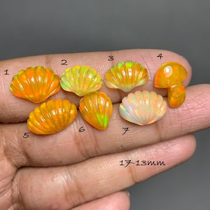 Puede incluir: Colección de siete piedras preciosas de ópalo talladas en formas de conchas y setas, que van del naranja al amarillo, con destellos iridiscentes. Los ópalos miden entre 17-13 mm. Las piedras se muestran en una mano.