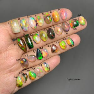 Peut inclure: Une main présente une collection d'opales de formes et de couleurs variées, notamment des coupes ovales, en goutte et rectangulaires. Les opales présentent un éventail de teintes, telles que le vert, l'orange et le violet, certaines affichant des motifs irisés. La taille des opales varie de 11 mm à 27 mm.