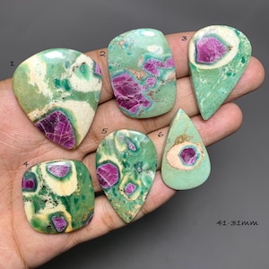 Puede incluir: Seis cabujones pulidos que presentan una mezcla de fuchsite verde e inclusiones de rubí magenta vibrante. Cada piedra está numerada del 1 al 6, mostrando patrones únicos de matriz crema, verde claro y verde oscuro. El tamaño es de 31-41 mm.