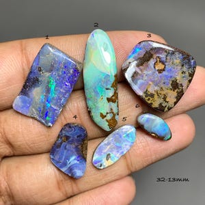 Puede incluir: Seis piedras preciosas de ópalo pulidas numeradas del 1 al 6, que muestran destellos iridiscentes de azul, verde y morado sobre roca matriz oscura. Las piedras varían en forma y tamaño, con el texto 32-13mm visible en la esquina inferior derecha.