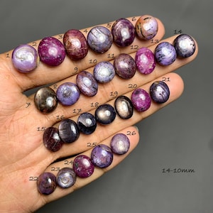 May include: A collection of twenty-six oval-shaped cabochon gemstones in shades of purple, pink, and dark brown, each marked with a black number from 1 to 26. The stones exhibit a star-like light reflection and range in size from 14 to 10 mm.