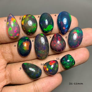 May include: Eleven polished black opals numbered 1 through 11, displaying iridescent flashes of red, green, and blue. The stones are oval, pear, and irregular shapes, ranging from 21-11mm in size. Each gem features a smooth, reflective surface.