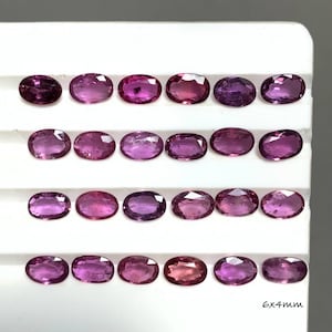 May include: A collection of twenty-four oval-cut, faceted magenta gemstones arranged in four rows on a white display tray. Each individual stone measures 6x4mm as indicated by the text printed on the bottom right of the display surface.