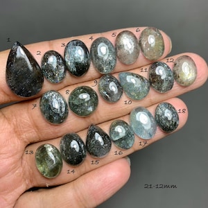 May include: Eighteen polished gemstones in diverse shapes, from teardrop to oval. The stones exhibit dark green, grey, and iridescent hues. The image includes the size range of 21-12mm. The gemstones are displayed on a hand.