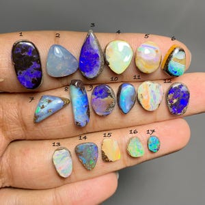 May include: Seventeen loose, polished opal gemstones arranged on skin, numbered 1 through 17. The stones display various shapes, including oval, teardrop, and triangular, with iridescent flashes of blue, purple, orange, green, and white colours.