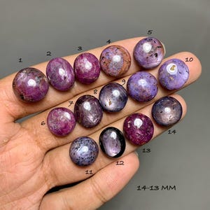May include: Fourteen polished, round and oval cabochon gemstones in shades of magenta, purple, and deep pink. Each stone is numbered from 1 to 14, displaying natural inclusions and reflective surfaces. The text 14-13 MM is visible at the bottom.
