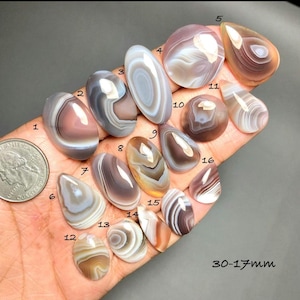 Puede incluir: Una colección de piedras preciosas de ágata pulidas en varias formas y colores, incluyendo marrón, blanco y gris. Las piedras preciosas miden entre 17 mm y 30 mm. El texto "30-17mm" es visible.
