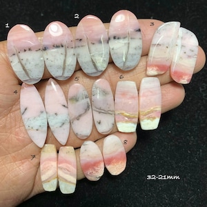 May include: A collection of eight polished gemstones in various shapes, including oval, teardrop, and rectangular. The stones display a mix of pink, white, and gray hues, with natural patterns. The text "32-21mm" is visible.