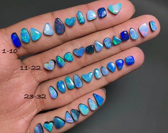 Puede incluir: Una colección de 40 piedras preciosas de ópalo en varias formas y tamaños, que van de 15 a 8 mm. Los ópalos muestran un espectro de colores, incluyendo azules, verdes y toques de rojo, con algunas piedras talladas en forma de corazón. Las piedras preciosas están dispuestas en una mano.