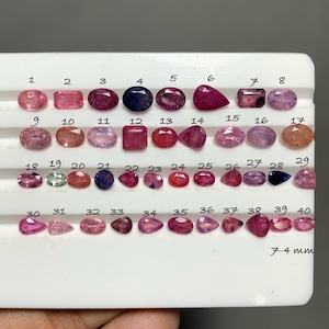 May include: A white display tray holding forty faceted loose gemstones in various shades of pink, red, and purple. Each stone is numbered from 1 to 40, featuring oval, pear, and rectangular cuts. The text 7-4 mm is printed in the bottom right corner.