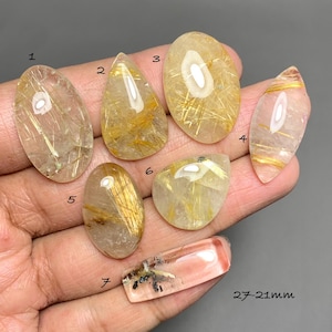 May include: Seven polished rutilated quartz cabochons with golden needle-like inclusions. The stones are numbered 1 through 7, with various shapes including oval, pear, and marquise. A rectangular piece labeled 7 measures 27-21mm in size.