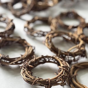 May include: A collection of small, circular woven grapevine wreaths. The wreaths are a natural brown color and appear to be handmade. They are arranged on a white surface, with some in focus and others blurred in the background.