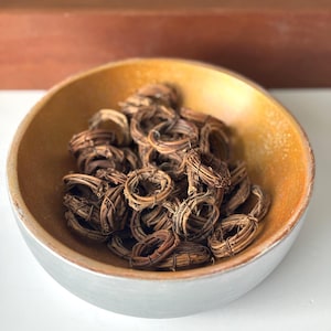 May include: A silver and gold-toned bowl filled with numerous small, woven, brown vine rings. The bowl has a metallic silver exterior and a golden interior. The rings are tightly wound and appear to be natural, organic elements.