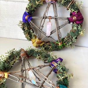 May include: Two rustic pentagram wreaths. Each wreath is crafted from twigs, adorned with green moss, colorful fabric accents, and wrapped with copper wire. One features a pink crystal pendant, the other a clear crystal.