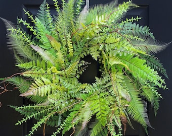 May include: A vibrant green fern wreath with a variety of textures and shades, hanging against a black door. The wreath features multiple types of ferns and brown vines, creating a natural and decorative accent for the home.