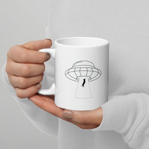 May include: White ceramic mug with a black line drawing of a cat being abducted by a UFO. The UFO is a classic flying saucer shape with a beam of light coming from the bottom.