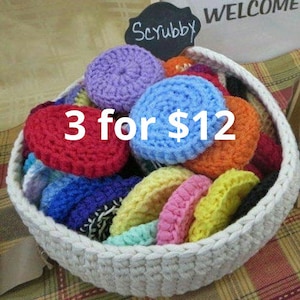 May include: A white crocheted basket filled with colorful crocheted scrubbies. The sign above reads "Scrubby" and "Welcome". The sign below reads "3 for $12".
