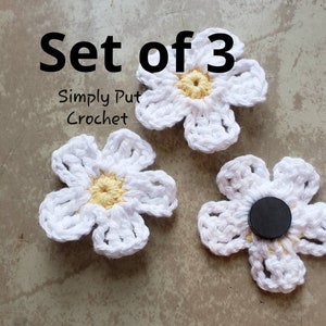 May include: Set of three white crocheted flowers with yellow centers. The flowers are on a gray background. The text "Set of 3" and "Simply Put Crochet" is visible in the image.