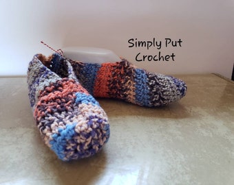 May include: A pair of crocheted slippers in shades of blue, pink, gray, and orange. The slippers are made with a textured stitch and have a simple, comfortable design. The text "Simply Put Crochet" is visible in the image.