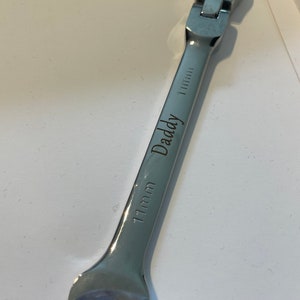 Op de afbeelding: Een zilveren sleutel met een ratelkop en een maatmarkering van 11 mm. De sleutel is gegraveerd met het woord "Daddy".