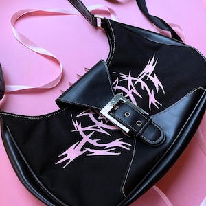 May include: Black shoulder bag with a pink tribal design. The bag has a black leather flap with a silver buckle closure. The bag has a black strap and white stitching. The background is pink.
