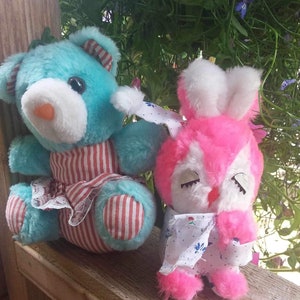 Può includere: Un orsacchiotto di peluche blu con un vestito a righe rosse e bianche e un coniglietto rosa di peluche con un vestito bianco con fiori blu. Entrambi gli animali di peluche sono seduti su una superficie di legno.