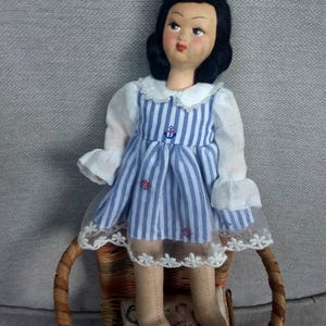 Op de afbeelding: Een porseleinen pop met zwart haar en een blauw-wit gestreepte jurk. De pop zit op een rieten stoel.