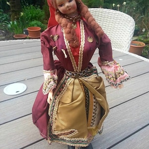 May include: A vintage doll in traditional costume. The doll is dressed in a burgundy dress with gold trim, a gold skirt, and a red headscarf. It has long brown plaits and is positioned on a wooden surface.