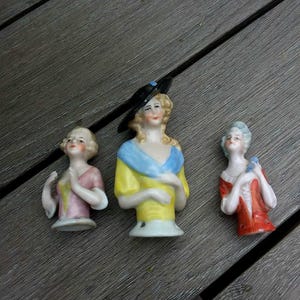 May include: Three vintage porcelain doll heads with painted faces and hair. The doll on the left is wearing a pink dress, the doll in the middle is wearing a yellow dress with a blue collar, and the doll on the right is wearing a red dress.