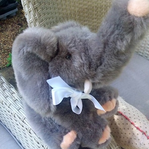 Può includere: Un giocattolo di elefante di peluche grigio con un fiocco bianco seduto su una sedia di vimini. L'elefante ha le orecchie rosa e una lunga proboscide.