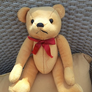 Può includere: Un orsacchiotto di peluche marrone con un papillon rosso seduto su un cuscino color crema. L'orso ha occhi neri e un naso nero.