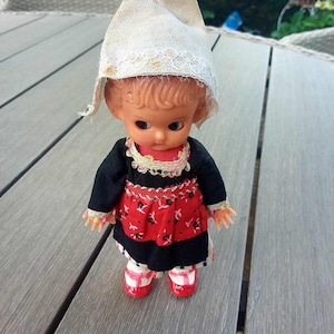 Op de afbeelding: Een kleine plastic pop met een witte muts, een zwarte jurk met rode en witte bies en rode schoenen. De pop staat op een houten oppervlak.