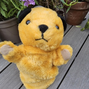 Op de afbeelding: Gele pluche pop met zwarte oren, een zwarte neus en mond. De pop heeft bruin gekleurde poten. De achtergrond bestaat uit planten en een houten terras.