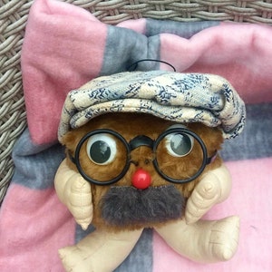 Może przedstawiać: Kapryśna pluszowa zabawka z brązowym futrzanym korpusem, dużymi oczami i czerwonym nosem. Nosi okrągłe okulary, szare wąsy i czapkę w wzory. Zabawka spoczywa na różowo-szarym pasiastym kocu.