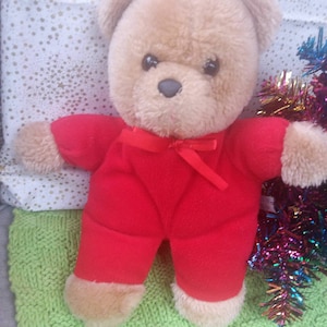 May include: A brown teddy bear wearing a red jumpsuit with a red ribbon around its neck. The bear is sitting on a green knitted surface with colourful tinsel in the background.