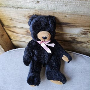 Peut inclure: Un ours en peluche noir avec un museau et des pattes beiges, et un ruban rose autour du cou. Le jouet en peluche est assis sur une surface gris clair, avec un fond en bois.