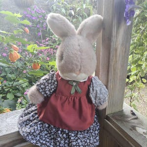 Op de afbeelding: Een pluche konijnenpop met lange oren, gekleed in een rode jurk met een rok en mouwen met bloemenpatroon. De pop staat buiten op een houten oppervlak, met een tuin op de achtergrond.