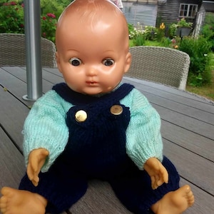 Puede incluir: Una muñeca de bebé vintage con ojos marrones y un atuendo de punto. La muñeca lleva un suéter azul claro y un mono azul oscuro con botones dorados. La muñeca está sentada sobre una superficie de madera.