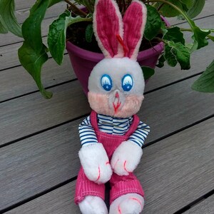 Op de afbeelding: Een wit pluche konijn dat een roze overall en een blauw-wit gestreept shirt draagt. Het konijn heeft roze oren en grote blauwe ogen.