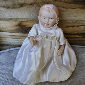 May include: A porcelain baby doll in a white satin dress and a pink bonnet. The dress features a delicate floral and pearl trim. The doll has blue eyes and a serene expression, a classic collectible item.
