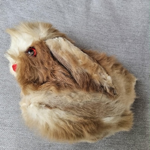 Può includere: Un giocattolo vintage a forma di coniglio in pelliccia marrone e beige, con occhi rossi e naso rosso. Il giocattolo ha lunghe orecchie ed è appoggiato su una superficie grigia. La pelliccia del coniglio sembra morbida e soffice.