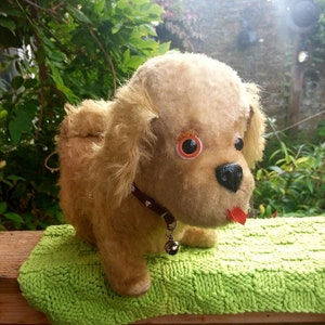 May include: A plush toy dog with brown fur, large brown eyes, and a red tongue sticking out. The dog is wearing a black studded collar with a silver bell.