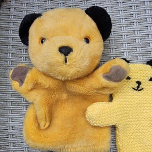 Può includere: Due burattini a forma di orso. Il primo è giallo con orecchie nere, naso e zampe marroni. Il secondo è giallo lavorato a maglia con un viso cucito nero.