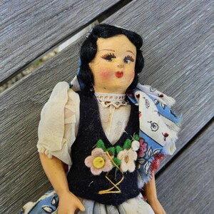 Könnte beinhalten: Eine Vintage-Puppe in traditioneller Kleidung. Die Puppe hat schwarzes Haar, rosige Wangen und rote Lippen. Sie trägt eine dunkelblaue Weste mit floraler Stickerei über einer weißen Bluse, einem gemusterten Schal und einem floralen Rock. Die Puppe ist etwa 20 cm groß.