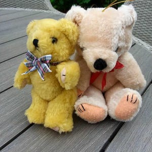Puede incluir: Dos osos de peluche, uno amarillo y otro crema, sentados en una mesa de madera gris. El oso amarillo tiene una corbata de lazo de cuadros rojos y negros. El oso crema tiene una corbata de lazo roja.