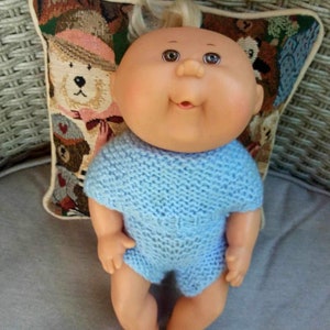Peut inclure: Une poupée Cabbage Patch Kid portant une tenue en tricot bleu clair. La poupée a les cheveux blonds et est assise sur un coussin à motif d'ours en peluche.