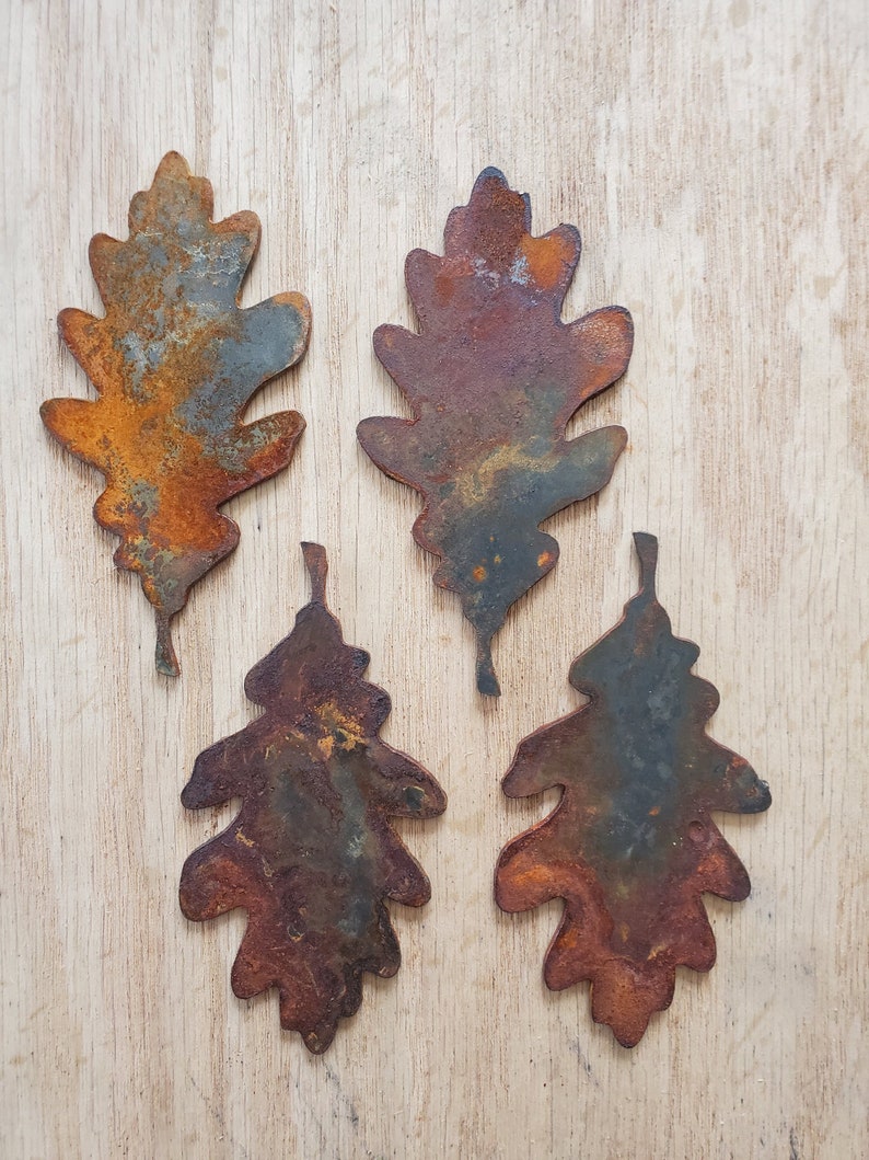 May include: Four rusty metal oak leaf shapes, each with a unique pattern of rust and oxidation. The leaves are arranged in a row on a light wood surface.