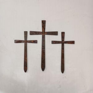 May include: Three rustic metal crosses of varying sizes are arranged against a white background. The crosses have a weathered, brown finish, suggesting an aged or antique appearance. The design is simple, with a horizontal bar intersecting a vertical one.